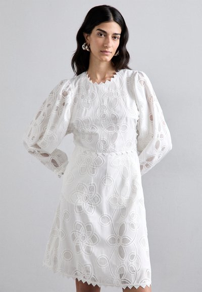 Robe blanche brodée présentant des motifs floraux, un col scallopé et des manches longues bouffantes. Tissu léger et texturé avec une taille cintrée.
