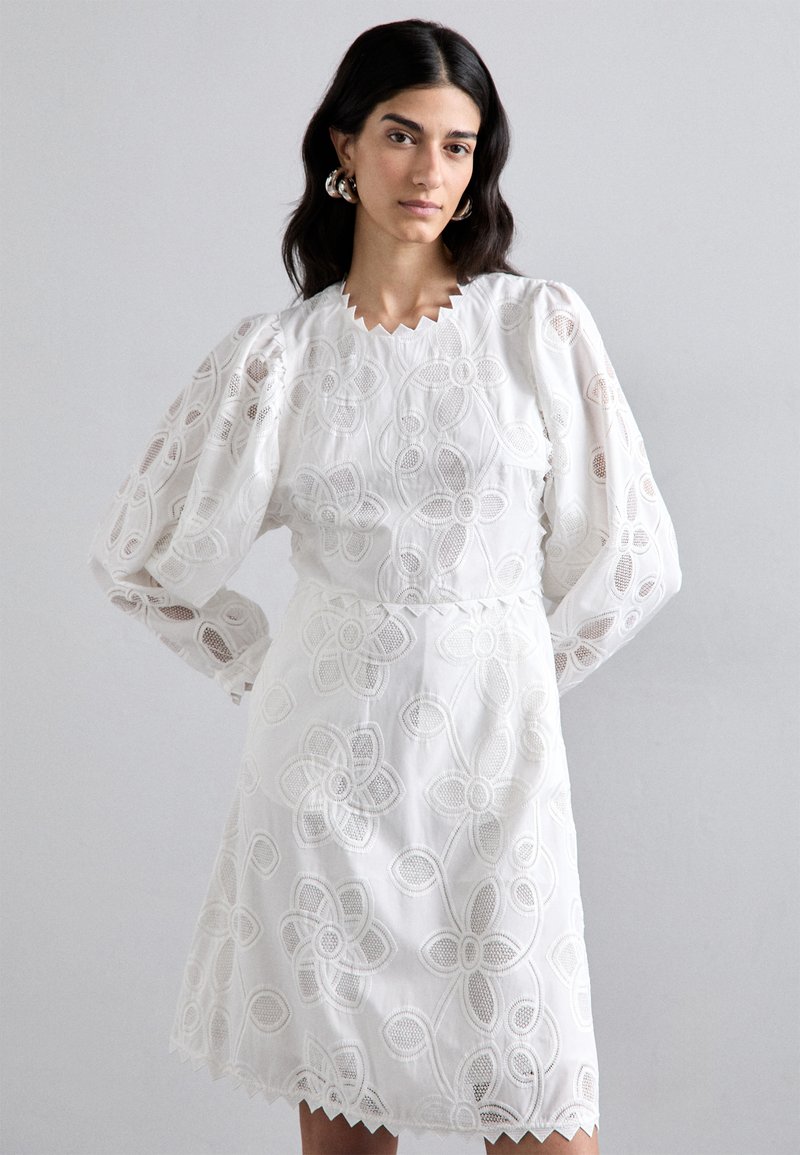 Robe blanche brodée présentant des motifs floraux, un col scallopé et des manches longues bouffantes. Tissu léger et texturé avec une taille cintrée.