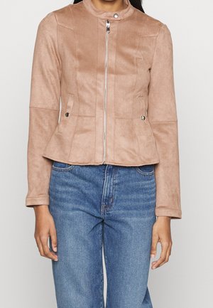 Kobieta w jasnobrązowej zamszowej kurtce z przednim zamkiem błyskawicznym i kołnierzem na zatrzaski, ubrana w wysokowycięte niebieskie dżinsy.