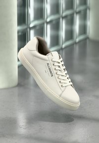 Λευκό αθλητικό παπούτσι AllSaints με κορδόνια, λογότυπο και ιαπωνικό κείμενο αιωρούμενο πάνω από ένα γυαλισμένο δάπεδο από σκυρόδεμα με θολωμένα γυάλινα τούβλα στο παρασκήνιο.