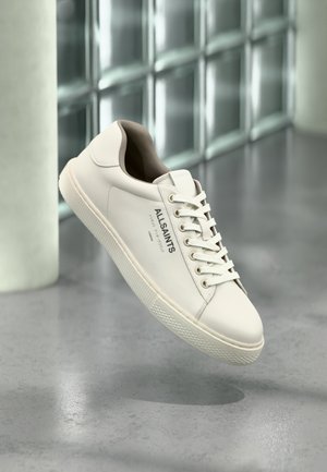 Biały sneaker AllSaints z sznurowadłami, logo i japońskim tekstem unoszący się nad polerowaną betonową podłogą z matowymi blokami szklanymi w tle.