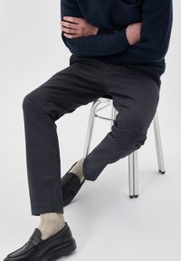Pantaloni grigi con texture e vestibilità slim, abbinati a mocassini neri e calzini beige chiaro, seduto su uno sgabello in cromo su uno sfondo bianco.