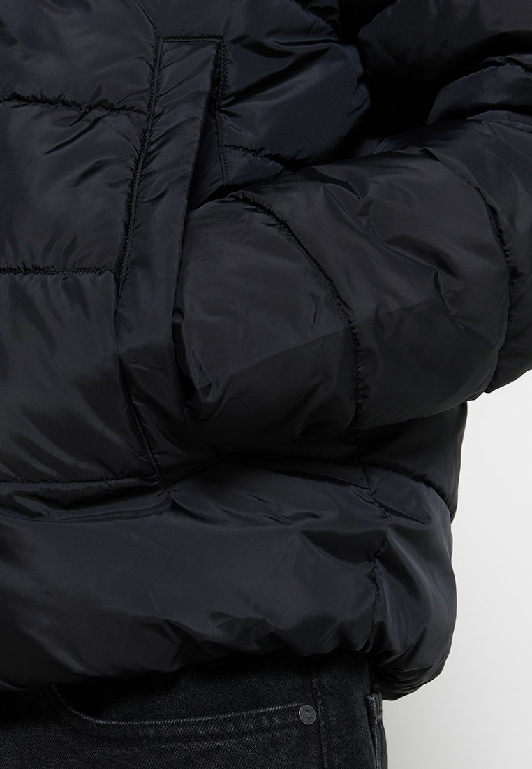 Chaqueta de plumas negra con diseño acolchado, que cuenta con un gran bolsillo lateral y una textura suave y brillante.