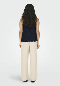 Marine blauwe mouwloze top met geschulpte onderrand en oogjesdetails, gecombineerd met lichtbeige wijde broek, gezien vanuit de achterkant.