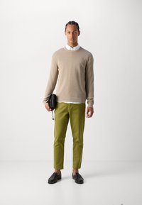 Pull en tricot beige, chemise blanche à col, chinos verts et chaussures noires. La personne tient un petit sac noir avec une bandoulière. Design simple et épuré.