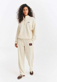 Béžový fleecový pulovr s polovičním zipem a vyšívaným logem, v kombinaci s odpovídajícími zúženými kalhotami s nášivkou loga a stahovacím pasem.