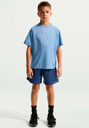 Niño de pie de frente con camiseta Nike azul claro, pantalones cortos Nike azul oscuro, calcetines Nike negros y zapatillas negras sobre fondo blanco.