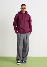 Sweat à capuche bordeaux avec une poche avant et un logo, associé à un pantalon large gris clair et des baskets noires avec des semelles blanches.