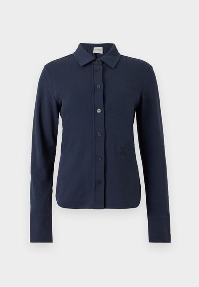 Chemise bleu marine à manches longues avec col, six boutons noirs sur le devant et un logo brodé discret sur le côté gauche.