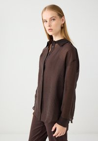 Camicia lunga trasparente marrone con colletto botton-down, texture sottile e vestibilità rilassata, abbinata a pantaloni scuri coordinati.