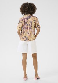 Camicetta a maniche corte con colletto, caratterizzata da un motivo astratto colorato in giallo, rosa e viola, abbinata a pantaloni corti bianchi e sandali con tacco cubano rosa.