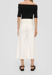 Una donna di spalle indossa una maglietta nera a spalle scoperte, pantaloni bianchi a vita alta e larghi, taglio cropped, e sandali neri con zeppa su sfondo grigio.