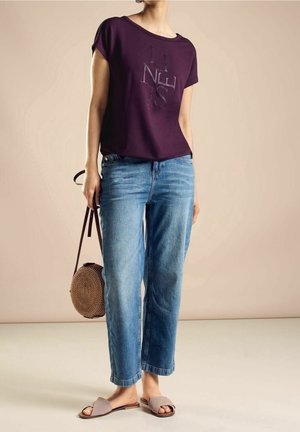 Henkilö, jolla on yllään väljä violetti grafiikkaprinttinen T-paita, siniset suoralahkeiset farkut, beiget liukusandaalit, ja joka pitää pyöreää pajukassia sivullaan.