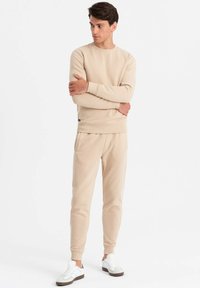 Beige Sweatshirt und passende Jogginghose in gerippter Textur. Rundhalsausschnitt und gebündelte Ärmel. Getragen mit weißen Turnschuhen mit braunen Akzenten.