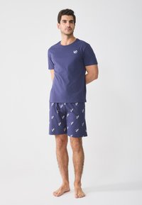 Marinblå bomulls-t-shirt med en liten logotyp på bröstet, kombinerad med mönstrade marinblå shorts som har små fågelmotiv. Modellen står barfota.