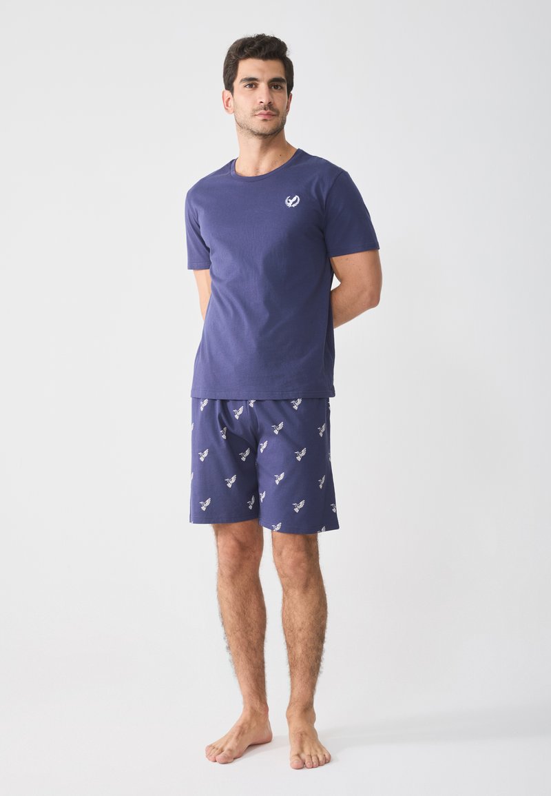 Marinblå bomulls-t-shirt med en liten logotyp på bröstet, kombinerad med mönstrade marinblå shorts som har små fågelmotiv. Modellen står barfota.