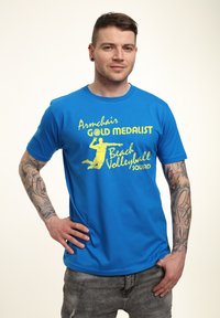 Camiseta de algodón azul con texto amarillo que dice "Equipo de Playa de Voleibol Armchair MEDALLISTA DE ORO", con un gráfico de una figura deportiva.