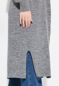 Grå, texturerad sweaterklänning med en sidoklyvning, kombinerad med mörkblå jeans. Har långa ärmar och en avslappnad passform.