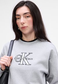 Γκρι φούτερ από βαμβάκι με στρογγυλή λαιμόκοψη. Διαθέτει κεντητό λογότυπο "Calvin Klein" σε μαύρο χρώμα. Το μοντέλο κρατά μια μαύρη τσάντα.