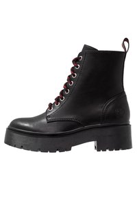 Bota de tobillo de cuero negro con una suela texturizada negra y gruesa. Cuenta con cordones rojos y ojales de metal, parte superior suave y una lengüeta en el talón.