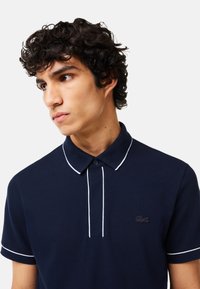 Marinblå polo med vit piping, korta ärmar och knäppt krage. Har en liten broderad logotyp på bröstet. Tyget är i bomull.