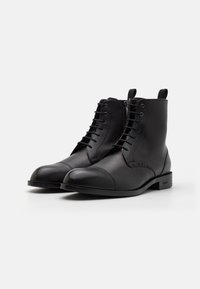 Botas de tobillo de cuero negro con un acabado liso, punta redondeada, cordones en la parte frontal y detalle de costura en contraste. Presentan una suela de perfil bajo.