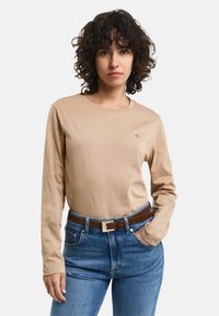 Frau mit lockigem Haar, die ein beiges Langarmhemd, eine blaue Jeans und einen braunen Gürtel trägt, steht mit einer Hand in ihrer Tasche.