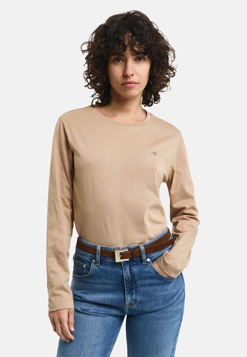 Frau mit lockigem Haar, die ein beiges Langarmhemd, eine blaue Jeans und einen braunen Gürtel trägt, steht mit einer Hand in ihrer Tasche.