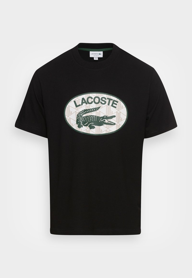 T-shirt noir à manches courtes avec un col rond, présentant un logo ovale vert et blanc avec un crocodile et le texte "LACOSTE" sur la poitrine.