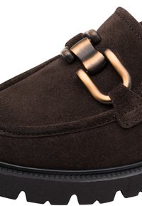 Mocassin en daim marron foncé avec une semelle épaisse en caoutchouc noir et un détail de boucle décorative en métal couleur bronze sur la tige.