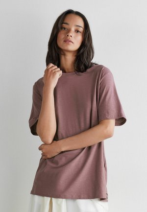 Vrouw met donker haar draagt een mauve T-shirt met korte mouwen en witte broek, staand tegen een effen lichtgrijze muur.