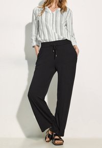 Pantalons larges noirs avec une taille élastique et un cordon de serrage, associés à une chemise à boutons rayée légère. Des sandales complètent le look.