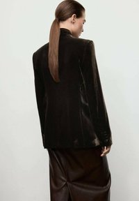 Mujer con una larga coleta marrón lleva un blazer de terciopelo oscuro y una falda de cuero oscura, de pie con su espalda hacia la cámara.