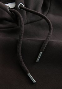 Brązowy materiał bluzy z kapturem z grubym, teksturowanym wykończeniem, z plecionymi sznurkami i metalowymi końcówkami. Zbliżenie podkreślające szczegóły.