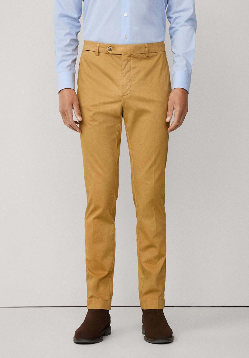 Uomo che indossa pantaloni slim-fit color senape, camicia dress azzurra chiara e stivali alla caviglia marrone scuro, in piedi contro uno sfondo chiaro e uniforme.