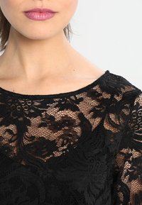Top in pizzo nero con un motivo floreale, finitura testurizzata e scollo ampio. Il tessuto appare delicato con dettagli intricati.
