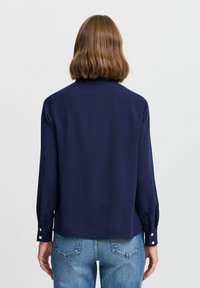 Blusa a manica lunga blu navy con una texture liscia, polsini abbottonati e una vestibilità rilassata, abbinata a jeans azzurri.