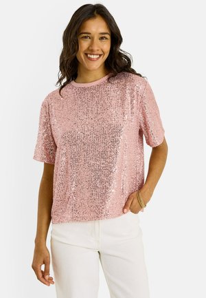 Mujer sonriente con cabello ondulado, vistiendo una camiseta rosa de manga corta con lentejuelas y pantalones blancos, de pie con una mano en el bolsillo.