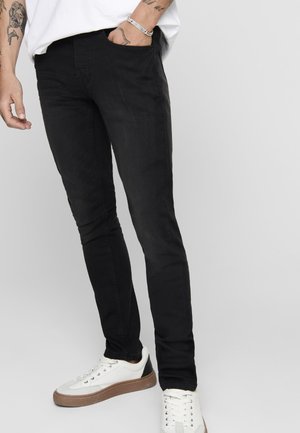Jeans negros de corte slim con una textura suave, que cuentan con bolsillos laterales y un diseño sencillo, combinados con zapatillas blancas con suela marrón.