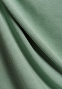 Tessuto verde chiaro con una texture liscia, adagiato per mostrare morbide pieghe e un leggero lucido. Nessun motivo o hardware distintivo visibile.