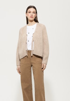 Beige knitted cardigan with tie closure, long sleeves, and ribbed cuffs; worn over a white top and paired with brown wide-leg trousers.