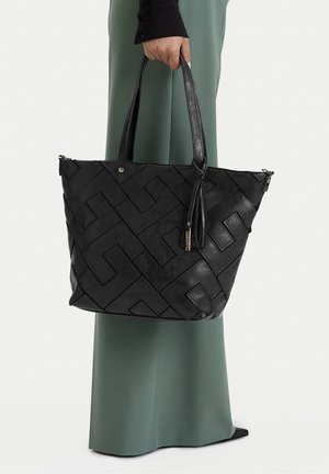 Persona sostiene un gran bolso tote de cuero negro con un patrón geométrico en relieve y detalle de borlas, vistiendo pantalones verdes de pierna ancha y una blusa negra.