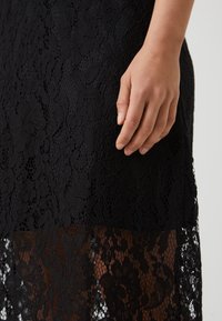 Black lace dress featuring a floral pattern with varying textures. The hem is sheer, revealing a layered lace effect. A hand is gently touching the fabric.