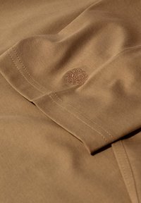 Brūns audums ar redzamām šuvēm un mazu izšūtu emblēmu, kas izceļ materiāla faktūru un darba meistarību.