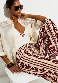 Mujer con gafas de sol, camisa blanca de encaje, top blanco y pantalones estampados con diseños florales color burdeos, sentada con un brazo detrás de la cabeza.