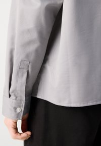Light gray long-sleeve shirt with a smooth texture, featuring buttoned cuffs and a curved hem, paired with black pants.