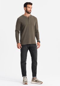 Camisa henley de manga larga verde oliva con botones, combinada con jeans gris oscuro y zapatillas grises con acentos beige. Diseño sencillo.