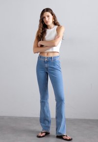 Jeans a zampa d'elefante azzurri chiari con vita alta, dotati di chiusura a bottoni e cinque tasche. Indossati con un top corto aderente bianco e infradito.