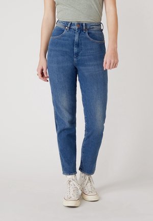 Vrouw met een blauwe high-waisted jeans, witte sneakers en een grijs aangesloten T-shirt, staand tegen een eenvoudige lichte achtergrond.
