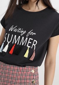 Černé bavlněné tričko s textem "Čekání na LÉTO", ozdobené barevnými střapci v červené, černé, žluté, modré a růžové barvě. V páru s kostkovanou sukní.